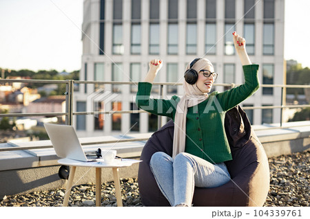 Woman in hijab hearing tunes via headphones on flat roof 104339761