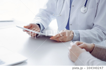 Doctor and patient sitting at the table in clinic while using tablet computer. The focus is on female patient's hands, close up. Medicine concept Doctor and patient sitting at the table in clinic while using tablet computer. The focus is on female patient's hands, close up. Medicine concept 104340247