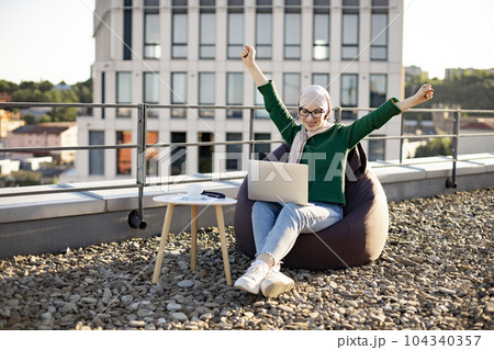 Female in hijab raising arms on rooftop with computer 104340357