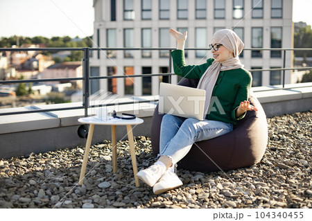 Lady in headscarf taking video call via laptop on rooftop Lady in headscarf taking video call via laptop on rooftop 104340455