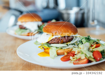 Closeup homemade burger with artificial meat, fried egg and vegetables on white plate with fresh salad on the kitchen table. Healthy fast food cooking recipe at home. Vegetarian diet. Selective focus. 104342192