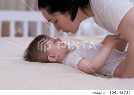 Loving mom leaning to kiss adorable little son lying on soft bed in nursery at home happy mother enjoying spending morning with chubby baby boy in white bodysuit 104342733