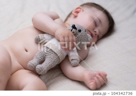 Cute chubby baby sleeping on back holding knitted bear toy with hand sweet little boy with closed eyes taking nap in comfortable bed in nursery at home Cute chubby baby sleeping on back holding knitted bear toy with hand sweet little boy with closed eyes taking nap in comfortable bed in nursery at home 104342736