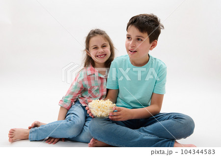 Mischievous adorable children, a teen boy and his younger sister sitting on white studio background, having fun together, eating popcorn and watching movie or cartoon. Kids and entertainment concept 104344583