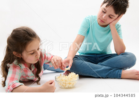 Caucasian happy primary school kids - teenager boy, brother and his younger sister, adorable litttle child girl eating popcorn, isolated on white studio background. Bonding. Family. Lifestyle Leisure 104344585