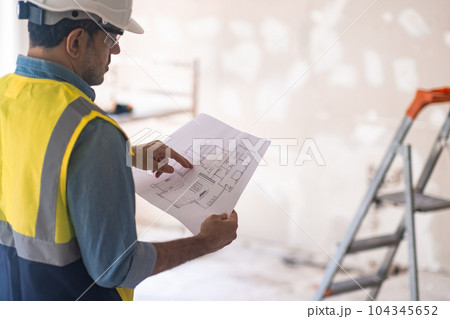 Concentrated worker in helmet checking apartment drawing stands in room in process of renovation experienced specialist in uniform standing in room against ladder near shabby wall 104345652