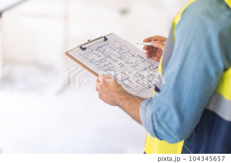 Engineer examining construction documentation holding clipboard and pen in hands man in professional uniform standing in unfinished premise ready for renovation 104345657