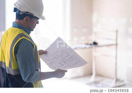 Focused architect with apartment drawing in hands and construction safety equipment man in protective helmet and vest reads details standing in light empty room 104345658