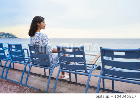 Beautiful smiling young mixed race woman sitting at one of the famous blue chairs at the Promenade des Anglais in Nice, France Beautiful smiling young mixed race woman sitting at one of the famous blue chairs at the Promenade des Anglais in Nice, France 104345817