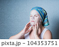 a girl in home clothes and with a towel on her head does a cosmetic procedure at home, using a sheet mask for facial skin care. 104353751