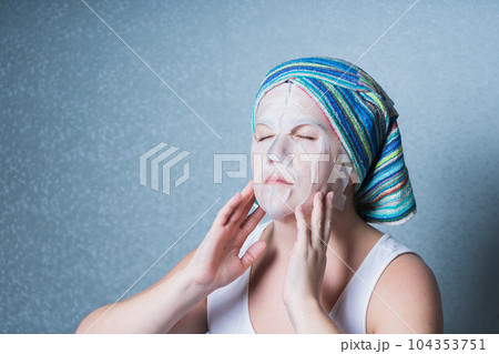 a girl in home clothes and with a towel on her head does a cosmetic procedure at home, using a sheet mask for facial skin care. 104353751
