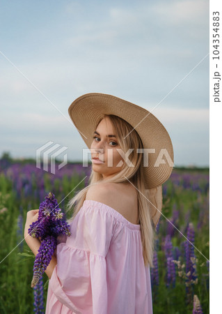 A beautiful woman in a straw hat walks in a field with purple flowers. A walk in nature in the lupin field 104354883
