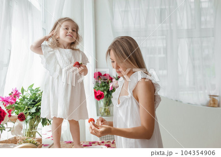 A little blonde girl with her mom on a kitchen countertop decorated with peonies. The concept of the relationship between mother and daughter. Spring atmosphere. 104354906