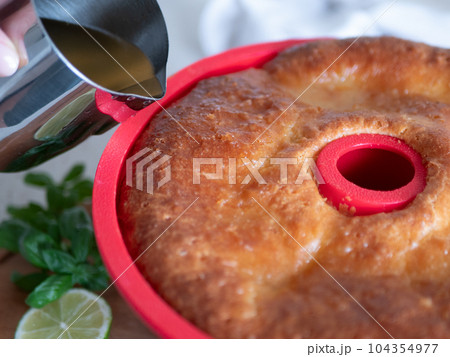 Pouring the syrup over freshly-baked rum baba in silicone cake mold. Rum-soaked circular cake Savarin Pouring the syrup over freshly-baked rum baba in silicone cake mold. Rum-soaked circular cake Savarin 104354977