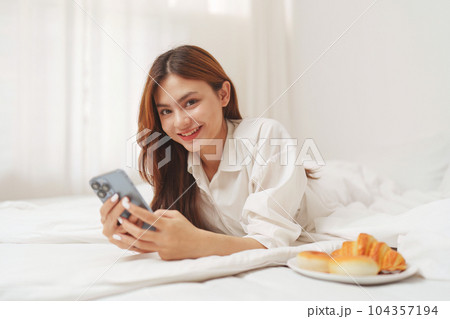 Happy smiling asian woman stretching on bed in the morning and holding mobile phone Happy smiling asian woman stretching on bed in the morning and holding mobile phone 104357194