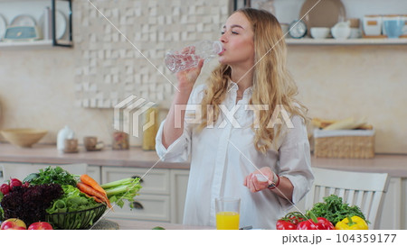 Beautiful woman standing in the kitchen and drinks mineral sparkling water from a plastic bottle. 104359177