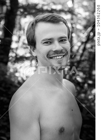 Black and white photo of Self assured young shirtless muscular male model with dark hair and beard smiling and looking at camera while standing in park . High quality photo 104362268