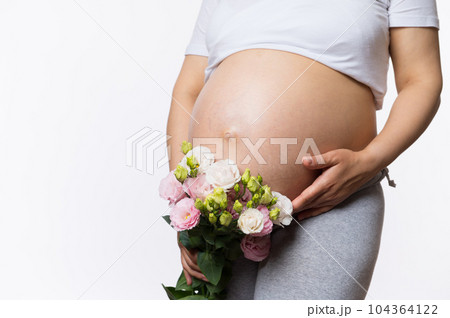 Beauty close-up portrait of gravid expectant mother holding bouquet of flowers near her pregnant belly, gently caressing her abdomen, isolated white background. Women's health and gynecology concept 104364122