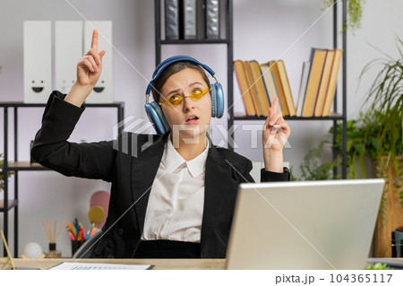 Caucasian businesswoman wearing headphones listening music dancing after working on laptop at office Caucasian businesswoman wearing headphones listening music dancing after working on laptop at office 104365117