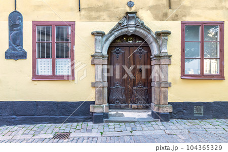 Decorated wooden door framed by stone frame, mediating wooden wrought iron windows in yellow wall and cobblestone floor 104365329