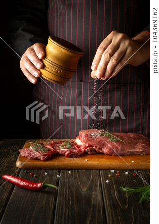 An experienced chef salts raw fresh beef before roasting or barbecuing. Working environment in the kitchen of a restaurant or hotel. 104369162