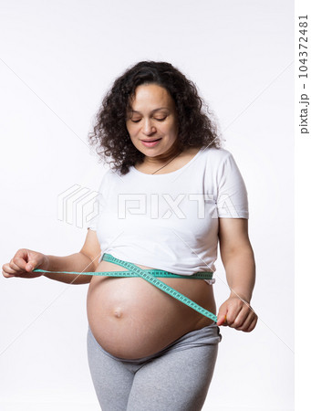 Advertising studio portrait of a curly haired adult pregnant woman, expectant mother, monitoring the development of her baby in womb, measuring her big belly with a tape, isolated on white background Advertising studio portrait of a curly haired adult pregnant woman, expectant mother, monitoring the development of her baby in womb, measuring her big belly with a tape, isolated on white background 104372481