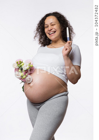 Smiling happy pregnant woman showing at camera a positive pregnancy test, posing with bunch of flowers, isolated over white background. Human fertility. Gynecology. Female health and maternity concept 104372482