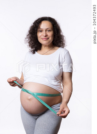Beautiful gravid pregnant woman, expectant mother measuring her big belly in third trimester with tape, isolated white studio background. Pregnancy. Maternity. Female health and gynecology concept 104372493