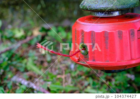 Trimmer reel with string, lawnmower on grass background. Red trimmer head 104374427