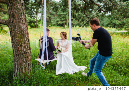 Videographer shoots a film with the newlyweds on a rope swing in a pine forest Videographer shoots a film with the newlyweds on a rope swing in a pine forest 104375447