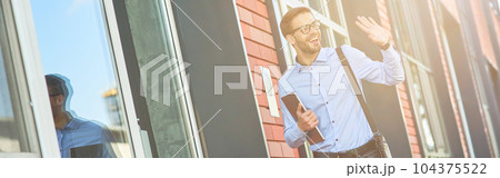 Young happy caucasian businessman wearing blue shirt and eyeglasses holding his laptop and waving, saying hello while standing against office building outdoors Young happy caucasian businessman wearing blue shirt and eyeglasses holding his laptop and waving, saying hello while standing against office building outdoors 104375522