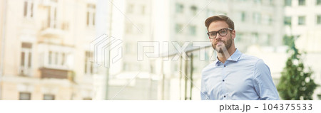 Young handsome caucasian businessman with suitcase waiting for taxi outdoors, standing on city street and looking away 104375533