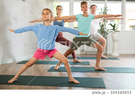 Pleasant little girl practicing warrior pose of yoga with her family 104380855