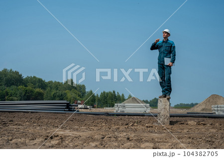 Builder in a protective helmet holds a laptop and stands on a pile in the middle of a construction site. Builder in a protective helmet holds a laptop and stands on a pile in the middle of a construction site. 104382705
