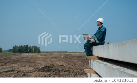 Caucasian male builder in hardhat sits on floor slabs and uses laptop at construction site. Caucasian male builder in hardhat sits on floor slabs and uses laptop at construction site. 104383013