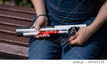 A blind woman sits on a bench and holds a folding tactile cane and sunglasses in her hands. A blind woman sits on a bench and holds a folding tactile cane and sunglasses in her hands. 104383136