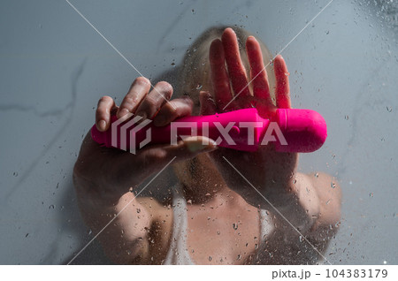A woman in a white tank top stands in the shower and holds a pink sex toy. A woman in a white tank top stands in the shower and holds a pink sex toy. 104383179