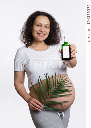 Smiling happy pregnant woman shows at camera a stretch marks massage oil with copy ad space, posing with palm leaf over white background. Natural vegan cosmetics, skin and body care, pregnancy concept Smiling happy pregnant woman shows at camera a stretch marks massage oil with copy ad space, posing with palm leaf over white background. Natural vegan cosmetics, skin and body care, pregnancy concept 104386376