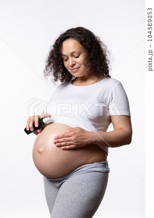 Vertical advertising studio portrait of expectant pregnant woman applying massage oil, moisturizing her big belly to avoid stretch marks during childbearing and pregnancy time, isolated white backdrop Vertical advertising studio portrait of expectant pregnant woman applying massage oil, moisturizing her big belly to avoid stretch marks during childbearing and pregnancy time, isolated white backdrop 104389853