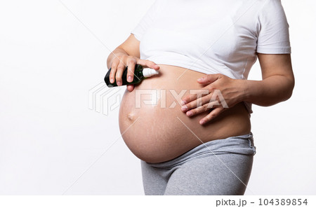 Close-up beautiful abdomen in pregnancy trimester third of a pregnant woman, applying natural smoothing cosmetic oil, massaging her big belly to avoid stretch marks, isolated white studio background Close-up beautiful abdomen in pregnancy trimester third of a pregnant woman, applying natural smoothing cosmetic oil, massaging her big belly to avoid stretch marks, isolated white studio background 104389854