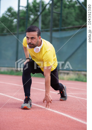 Man in yellow t-shirt doing forward lunge and touching ground with fingers at training on running track with rubber surface and white lines on urban sports arena 104392380