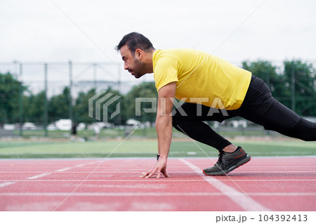 Concentrated athlete standing in low start pose before sprint on red rubberized track surface on city sports ground sportsman exercising at outdoor arena Concentrated athlete standing in low start pose before sprint on red rubberized track surface on city sports ground sportsman exercising at outdoor arena 104392413