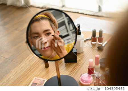 Reflecting in mirror of young gay man doing daily makeup in living room. LGBTQ, lifestyle and makeup concept Reflecting in mirror of young gay man doing daily makeup in living room. LGBTQ, lifestyle and makeup concept 104395483