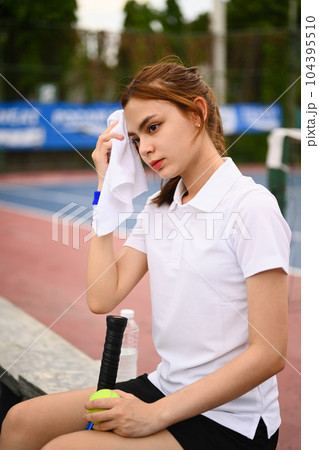 Pretty female tennis player wiping sweat with towel, resting on the bench at the tennis court after training Pretty female tennis player wiping sweat with towel, resting on the bench at the tennis court after training 104395510