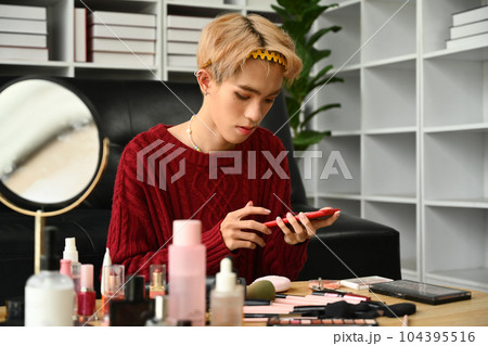 Young gay man doing daily makeup in living room with rainbow flag on background. LGBTQ, lifestyle and makeup concept 104395516