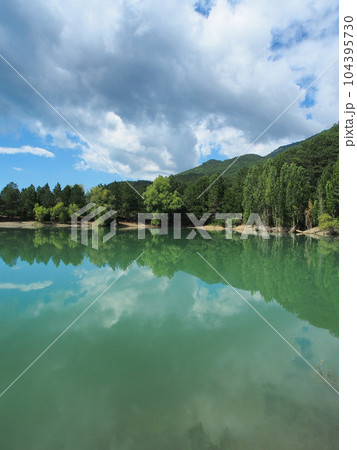 A summer landscape, a mountain lake surrounded by trees reflected in the water, against a blue sky with clouds. Mountain landscape, a beautiful place to relax. A summer landscape, a mountain lake surrounded by trees reflected in the water, against a blue sky with clouds. Mountain landscape, a beautiful place to relax. 104395730