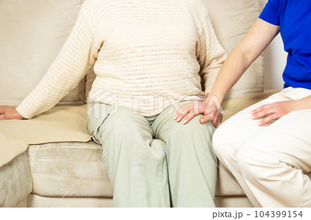 Young caregiver helping senior woman with knee pains at home. Caring nurse assisting her patient. Young caregiver helping senior woman with knee pains at home. Caring nurse assisting her patient. 104399154