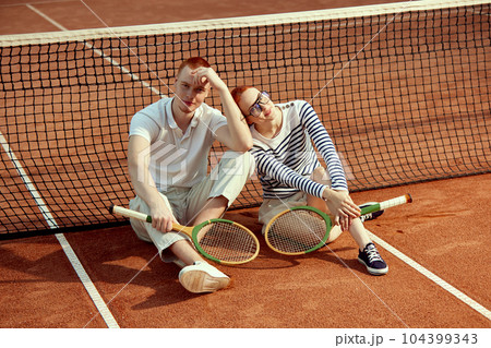 Handsome young man and beautiful woman in casual, stylish clothes posing with tennis racket on court on daytime Handsome young man and beautiful woman in casual, stylish clothes posing with tennis racket on court on daytime 104399343