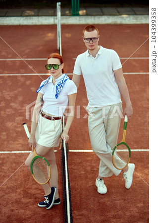 Full-length top view image of beautiful young woman and handsome man in casual, stylish clothes posing on tennis court on daytime Full-length top view image of beautiful young woman and handsome man in casual, stylish clothes posing on tennis court on daytime 104399358