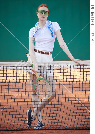 Full-length image of pretty, stylish, young redhead girl in casual clothes and trendy sunglasses posing with tennis racket on court Full-length image of pretty, stylish, young redhead girl in casual clothes and trendy sunglasses posing with tennis racket on court 104399361